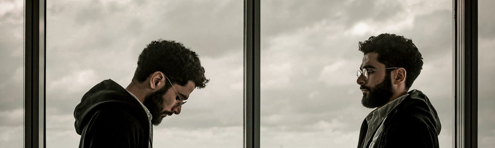 Adult male twins in a skyscraper overlooking a city. They appear depressed or disturbed in some way.