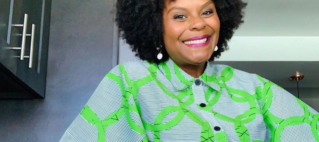 Tabitha Brown posing in a kitchen while wearing a bold graphic print dress.