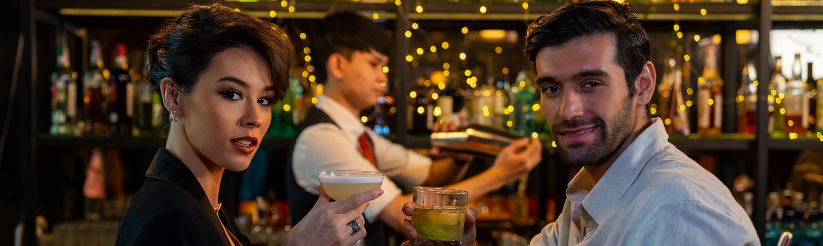 An attractive man and woman are sitting at a bar, looking directly at the viewer, seductively raising their cocktail glasses as if inviting the viewer to join them.
