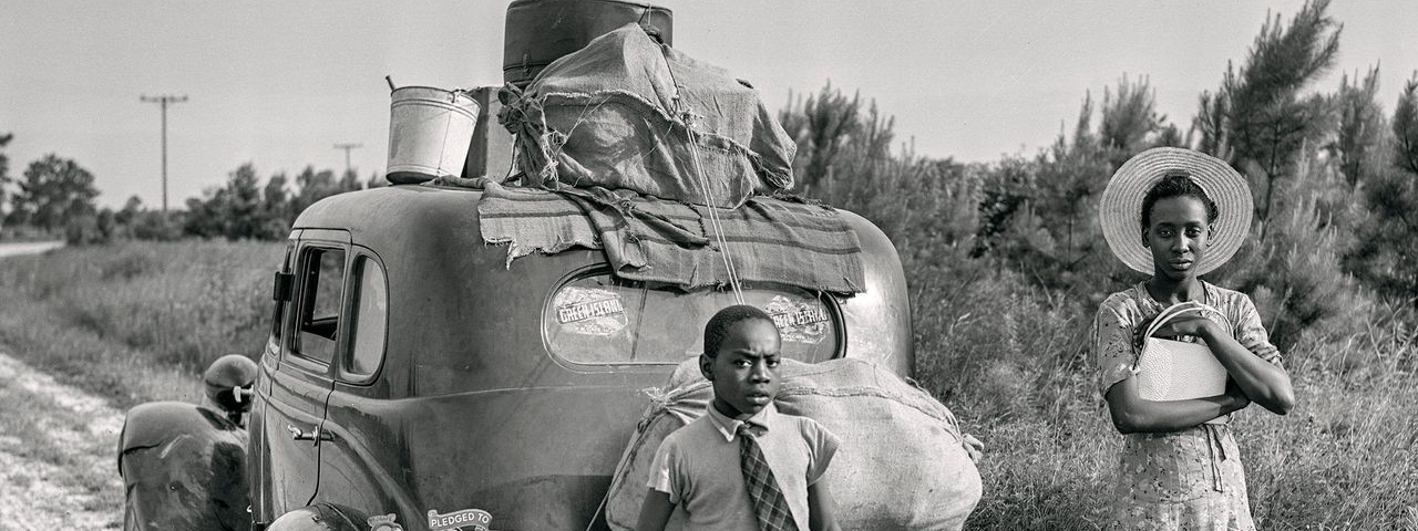 A black and white photo of two Black children standing in front of a 1940s car loaded with suitcases.