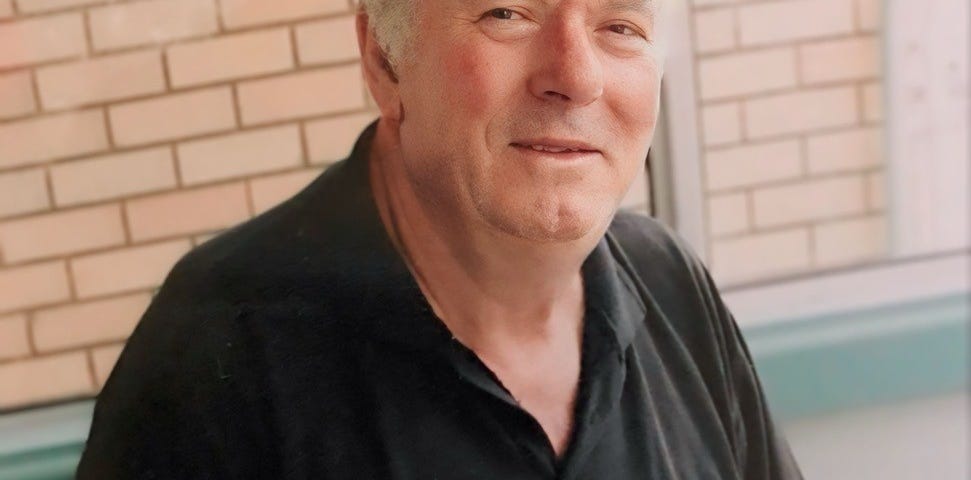 A man in his 70s with grey hair, in a black polo shirt, looking at the camera and smiling slightly.