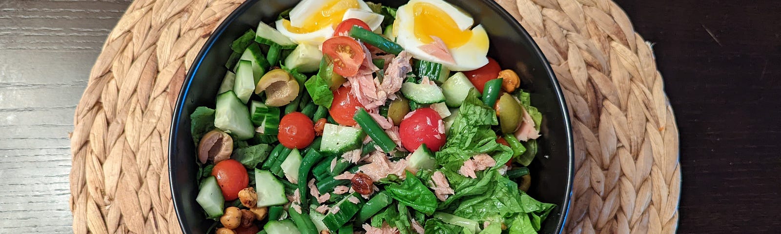 A salad is in a black bowl on a wicker placemat. The salad has greens, tomatoes, eggs, cucumber, chickpeas, olives, and tuna