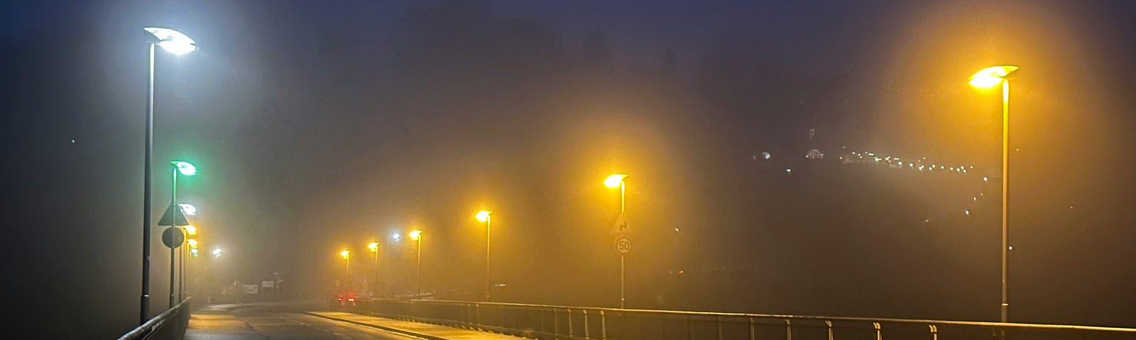 A deserted, foggy, wet road looks ominous at night with the only sign of life being the amber and white street lights illuminating the road.