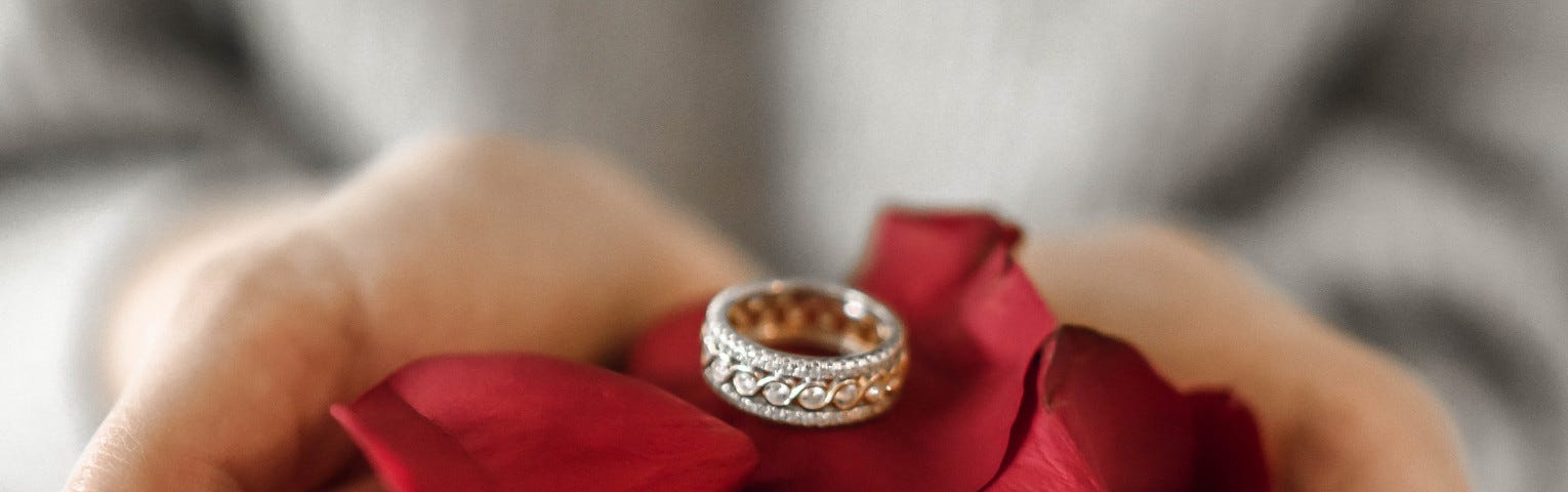 A man’s hands, offering a ring resting on rose petals.