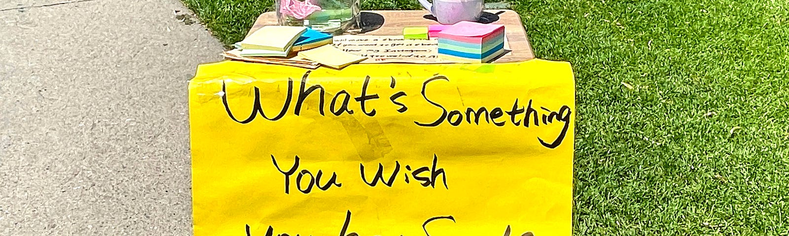 Small table with a bright yellow handwritten sign on front saying “What’s something you wish you have said?” glass jar on table top with a few pieces of paper. stack of colored sticky note. White mug with pens, some with pompoms on top.