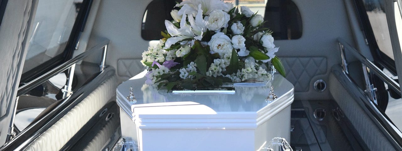 Image shows a white coffin in the back of a hearse, and on the coffin a bouquet with white flowers.