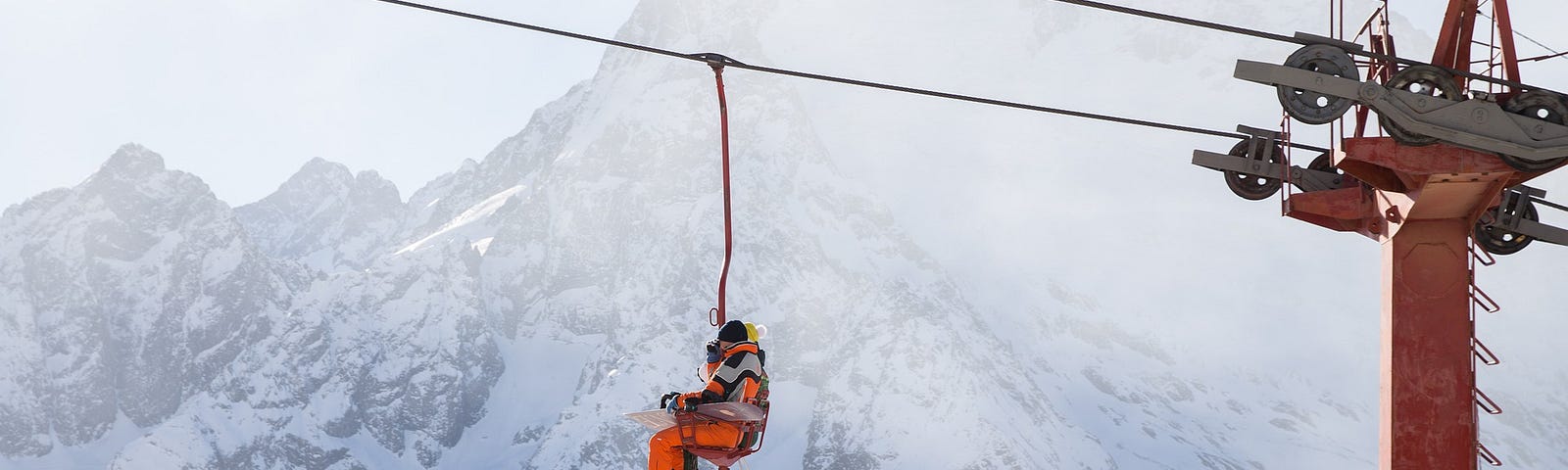 A skier riding up a chairlift with a snow-covered mountain in the background