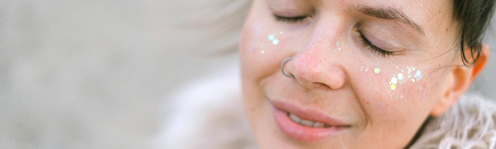 A young woman with glitter on her cheeks smiles gently with closed eyes