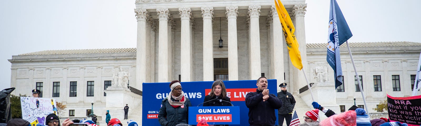 A crowd gathers in front of the Supreme Court for a rally. Signs and banners say, “gun laws save lives”