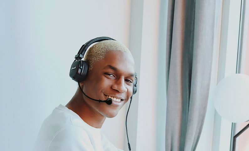 Image of young person, with close cropped hair, looking at camera and smiling brightly. He’s sitting at a laptop and is wearing a headset with a mic.