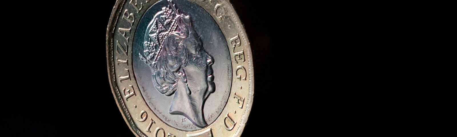 A coin balancing on its edge. The coin is a British pound coin and the  heads side is facing us. The heads side features Queen Elizabeth II’s profile.