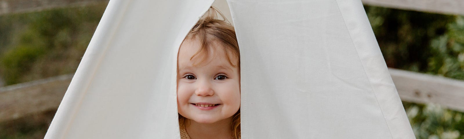 An angelic smiling baby girl peeking out naughtily from under a small white tent.