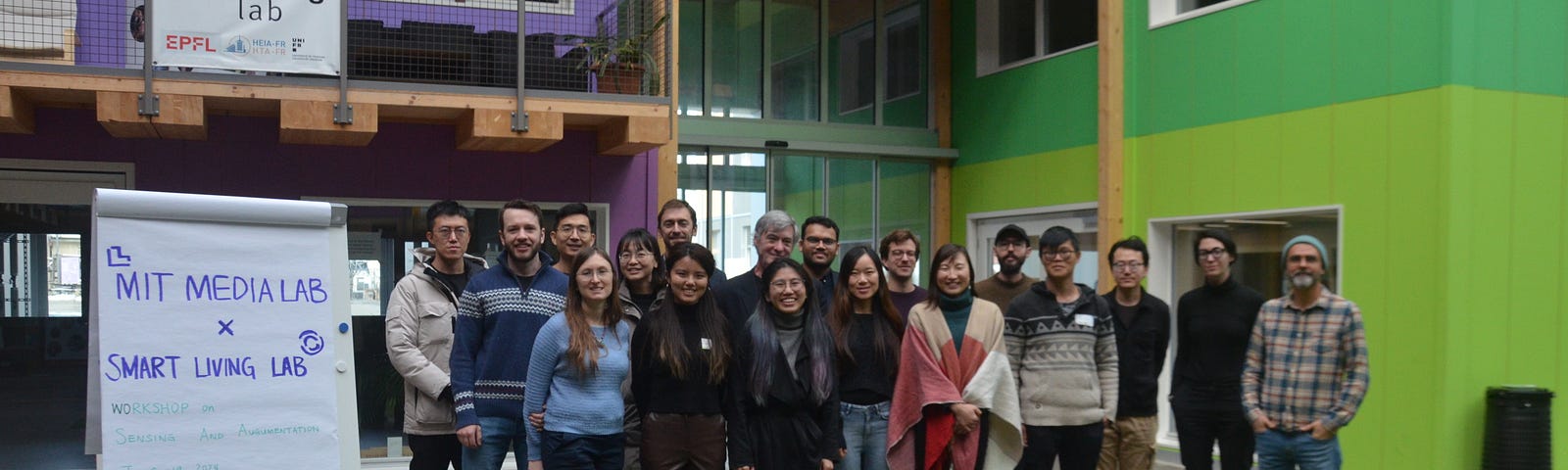 A group of people stand inside a building with green walls and turf flooring; a large white paper easel stands on the left side of the image with purple handwritten words on it saying “MIT Media Lab x Smart Living Lab.”