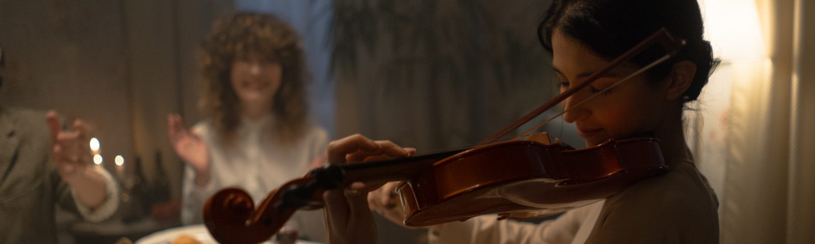 Woman playing a violin in a dimly lit room as background music for a dinner. There is another woman sitting at the dinner table in the background, looking like she is about to clap.