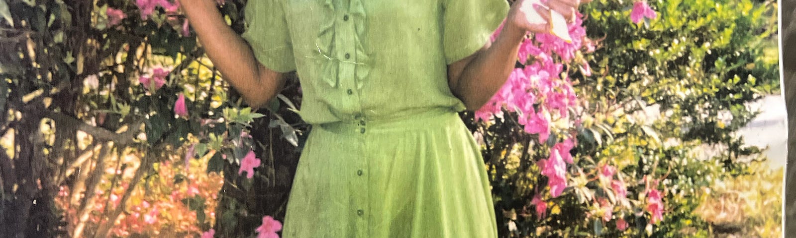 Authors mother, pictured in a bright green, linen, button down dress, standing in front pink flowers all around her.