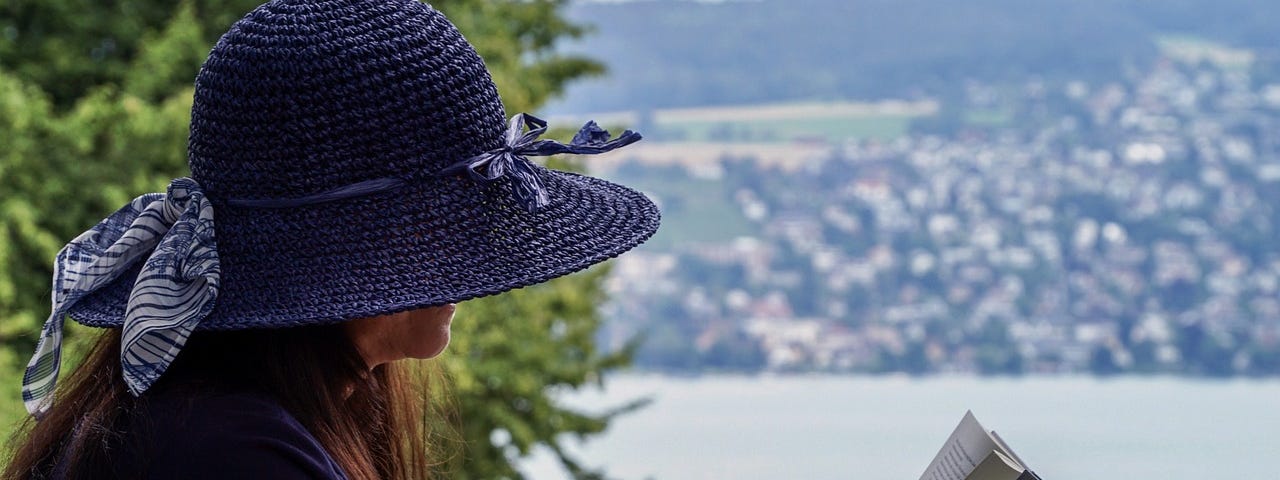 Woman With A Blue Straw Hat Sitting By A Lake Reading A Book.