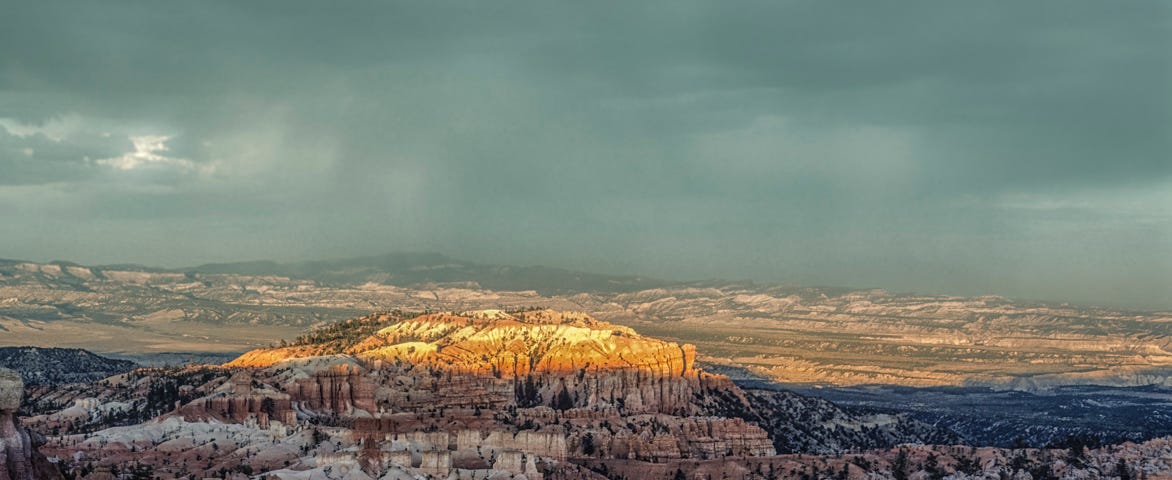 Dark cloudy sky with rain in background. Foreground mesa is almost covered with shadows. But the top of the mesa (close to center) is bathed in sunlight.