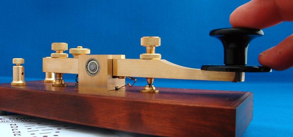 a morse key. it is a shiny brass metal piece mounted on a block of wood, fingers resting on it seeming ready to tap. it looks like new. underneath the key is a sheet of paper with the morse alphabet on it
