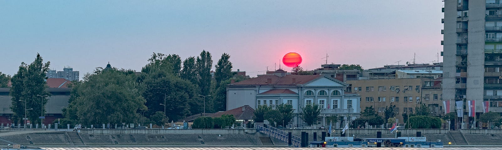 A bright red sun floats over historic buildings and a river. A boat floats on the river.