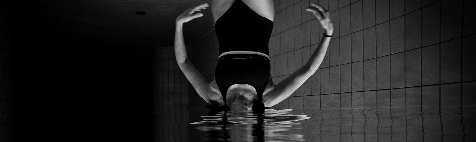 A black and white underwater image of girl in a black one-piece bathing suit in a pool, with the reflection obscuring her head such that her neck and the reflection of her neck meet at the water line. The image is rotated 180 degrees so that the reflection is the bottom, upright version of the girl.