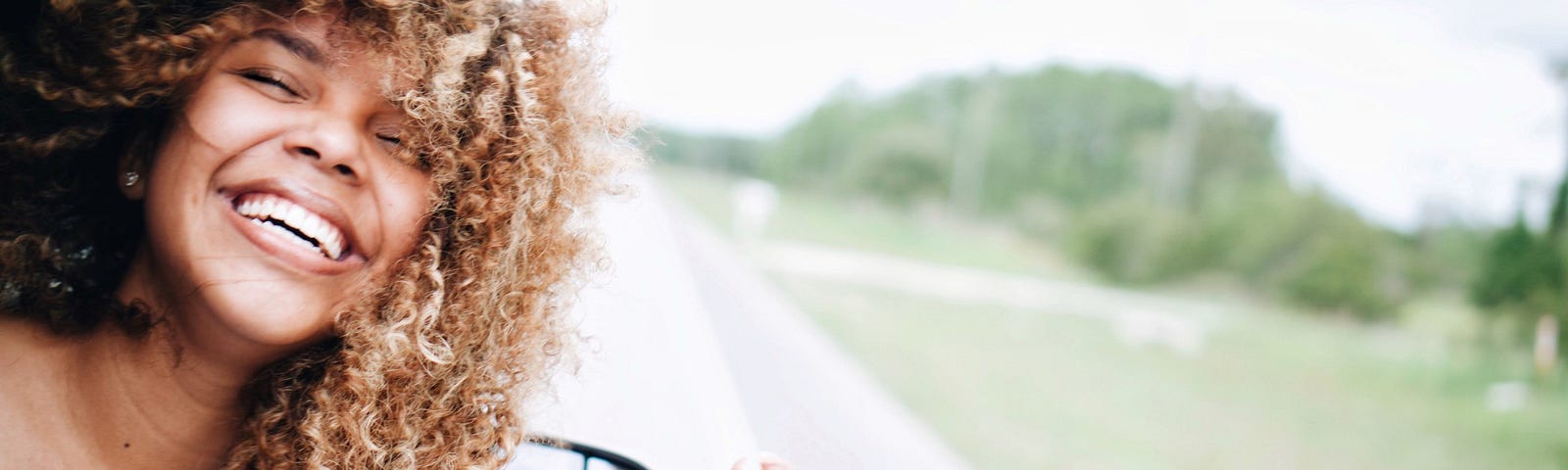 A black woman with natural hair smiles and leans from the window of a car.