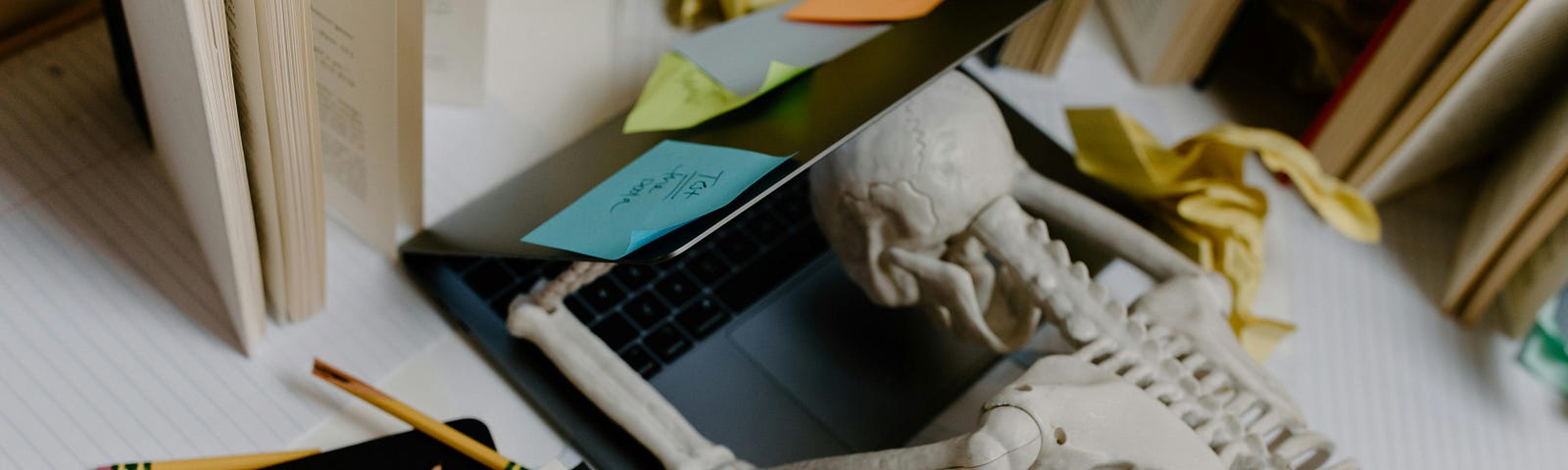 A plastic skeleton is collapsed into an open laptop on a cluttered workspace. The letters “S.O.S” are written on a notebook nearby.