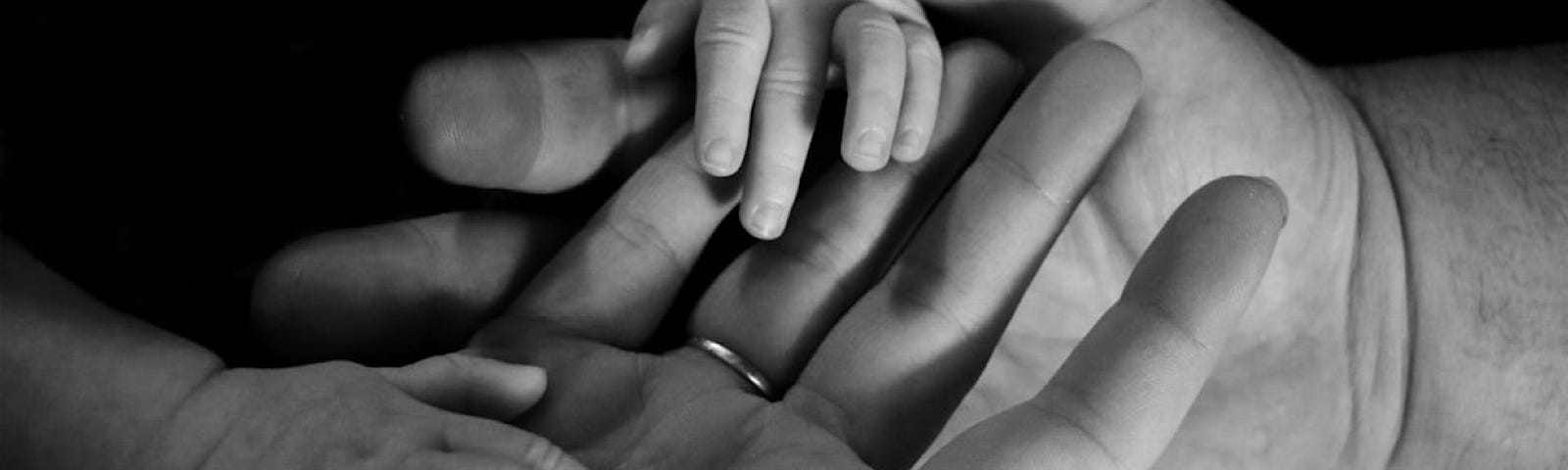 Black & White photo of an adult hand in a younger person’s hand, then two small hands of a baby resting on top. A collage of hands from all stages of life.