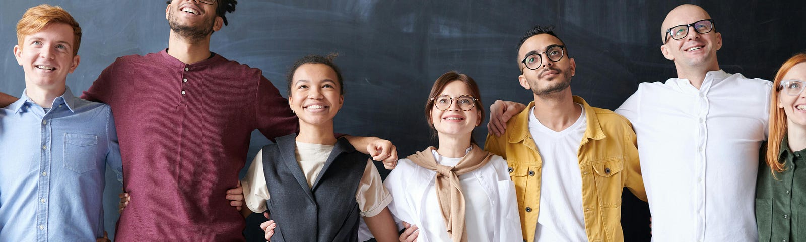 A team of people, wrapping arms around each other’s back, smiling in front of a classroom chalkboard.