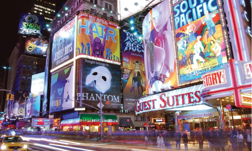 A bright city-scape street, many buildings are having posters for musicals such as “Wicked,” “Phantom of the Opera,” “Hair,” and others that are not readable. There are crowds of people walking down the streets, some entering the building with the sign “Guest Suites.” There is a taxi in the corner waiting and the entire picture is in bright and glowing colors.