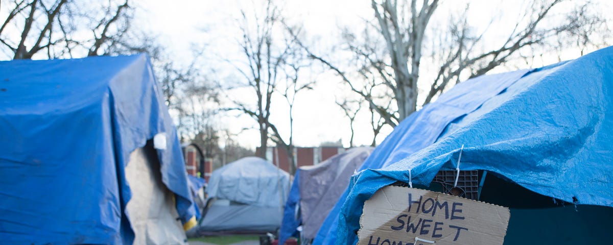 Image of a tent city with blue tarp covered tents. A tent in the foreground has a cardboard sign hanging from it