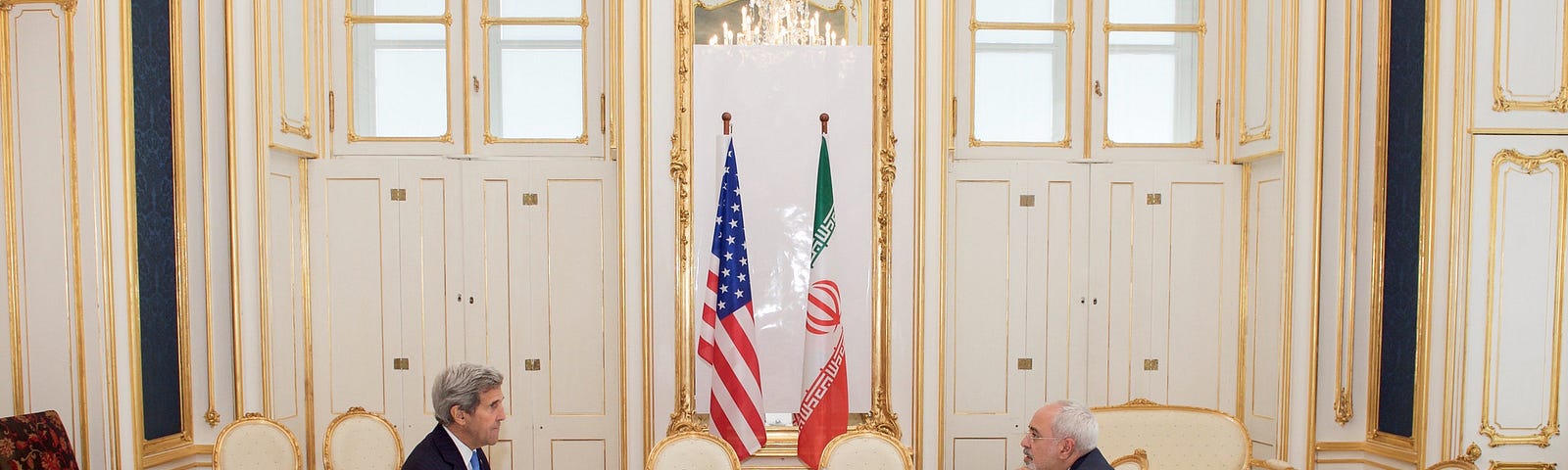 John Kerry and Javad Zarif sit across a table in front of American and Iranian flags in the Blue Salon of the Palais Coburg in Vienna.