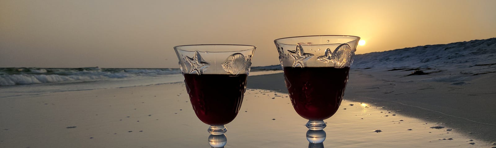 Two wine glasses half-filled with wine perched upon the shoreline of a beach at sunset.