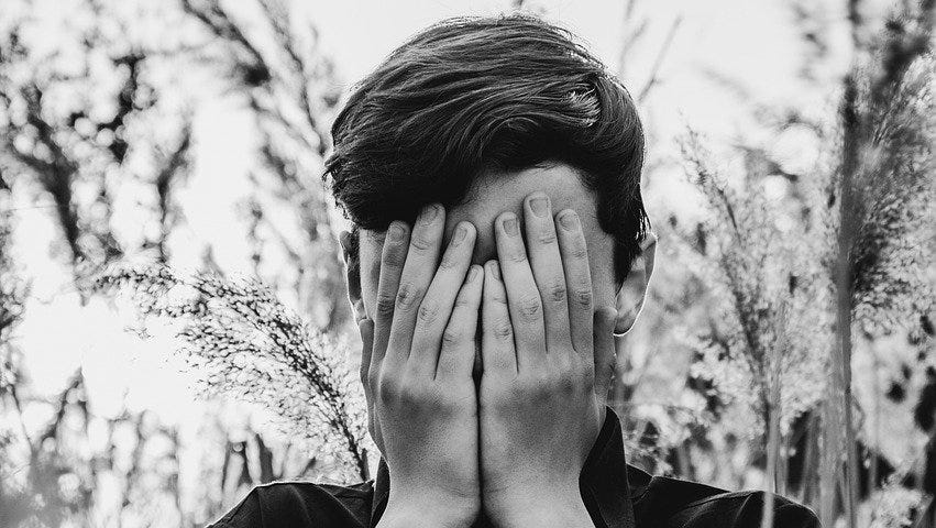 A man stands wearing a black turtleneck with his hands covering his face. He is against a black and white background of what looks like a winter forest.