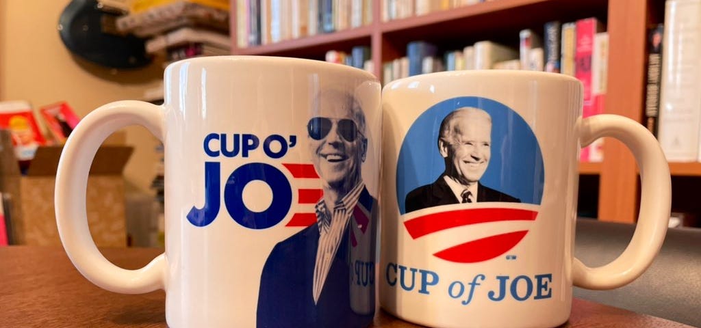 Two coffee mugs sitting on a dining table, one featuring a picture of Joe Biden inserted into the center of the Obama 2012 campaign logo with the text “Cup of Joe” written underneath, the other featuring a smiling portrait of Biden in his signature sunglasses, with the words “Cup o’ Joe” next to it.