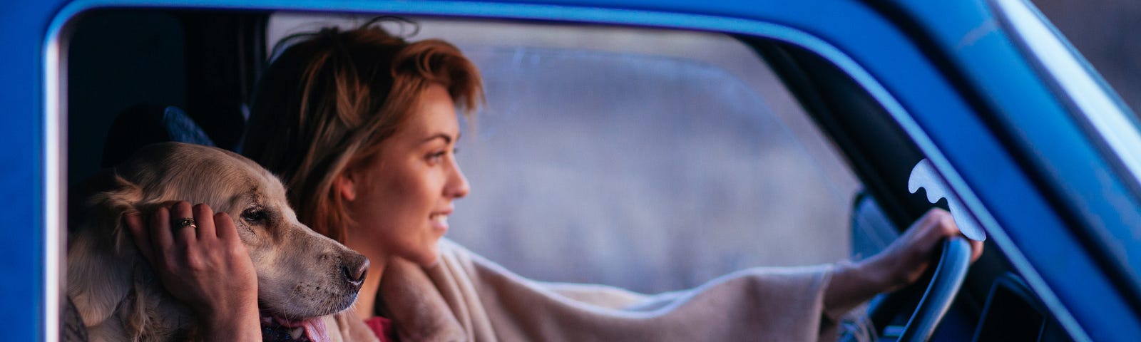 A woman in a blue truck smiles as she holds her steering wheel and pets her dog, whose head rests on her shoulder.