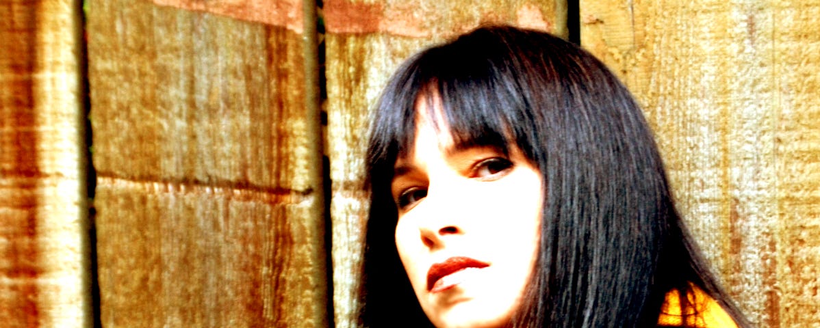 A woman with dark hair and bangs is looking at the camera in a 3/4 profile. She is standing in front of a wood fence and she’s wearing a brown leather jacket with a yellow scarf.