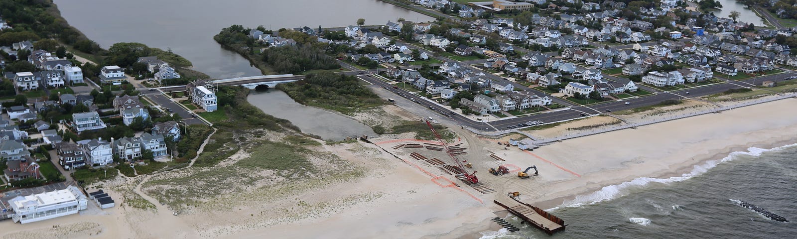 Aerial photo of coastal community. Beach separates the ocean from a waterway leading to a pond. Heavy machinery is working on the beach