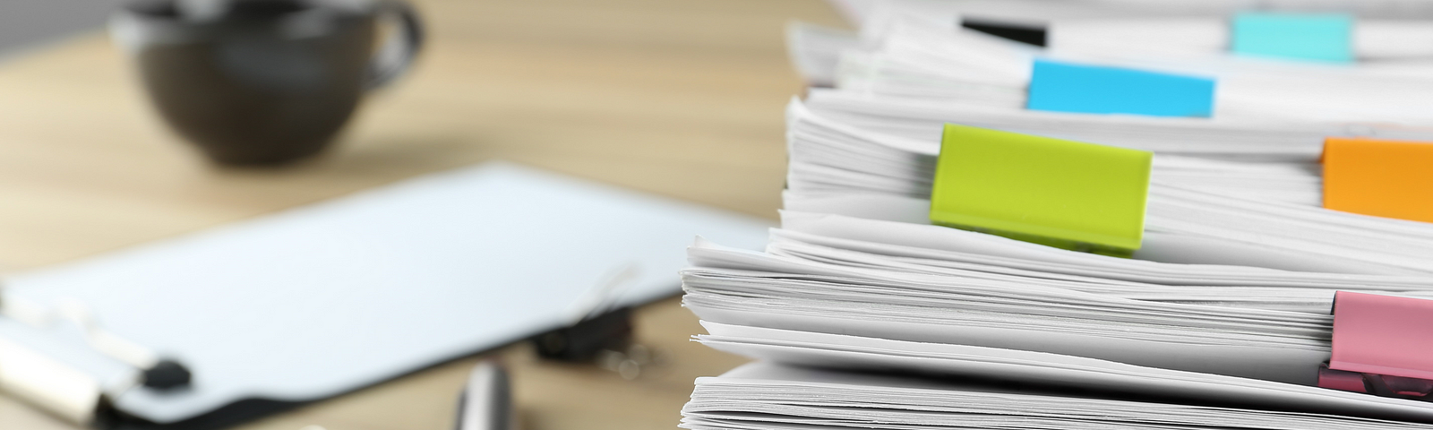 Stacks of papers held together with bulldog clips in the foreground. A pen, a clipboard and a coffee cup in the background.