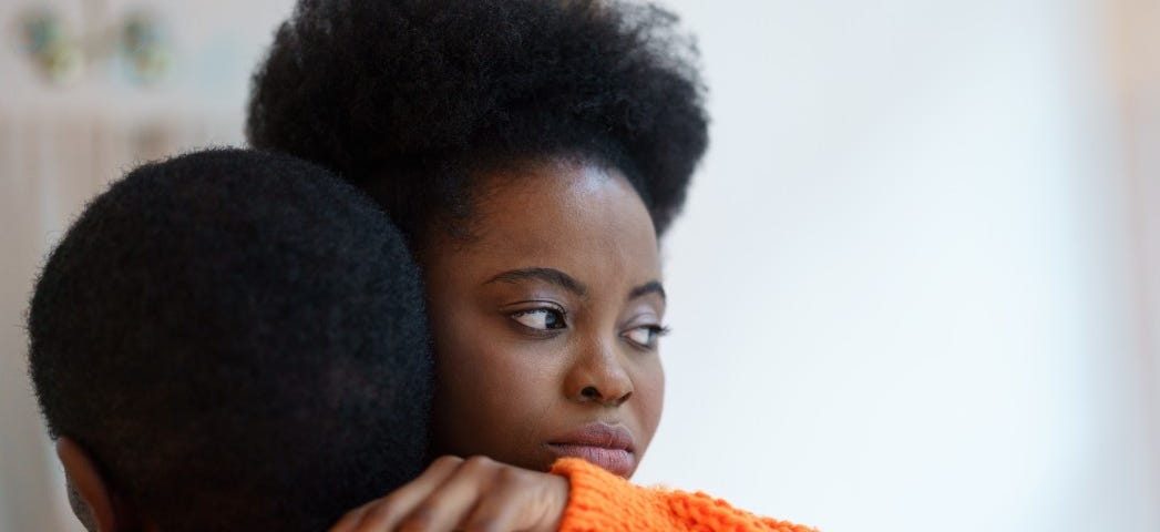 A woman in an orange sweater embraces her partner while gazing sadly away.