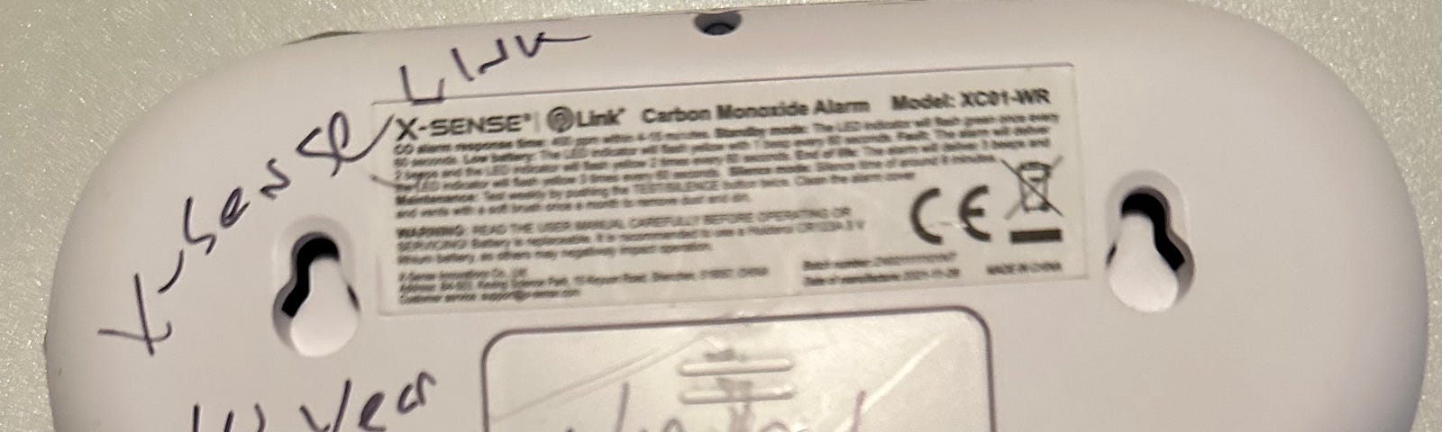 The image shows the back of a carbon monoxide alarm, specifically the X-Sense model XC01-WR. There is handwriting on the alarm that seems to indicate a lifespan or replacement schedule for the device, marked as “10 years”, with a date written below, “1/13/21”, which could possibly be the installation date or the date when the alarm was last checked. The presence of CE marking indicates that the product is compliant with European Economic Area standards for health, safety, and environmental prote