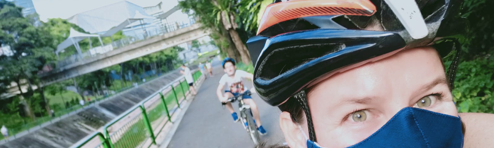 woman wearing a cycling helmet and face mask. She has green eyes and looks to be smiling under the mask. Behind her is an out of focus happy looking child on bicycle also wearing a helmet. They are on a cycle path with a bridge, buildings and trees in the background.