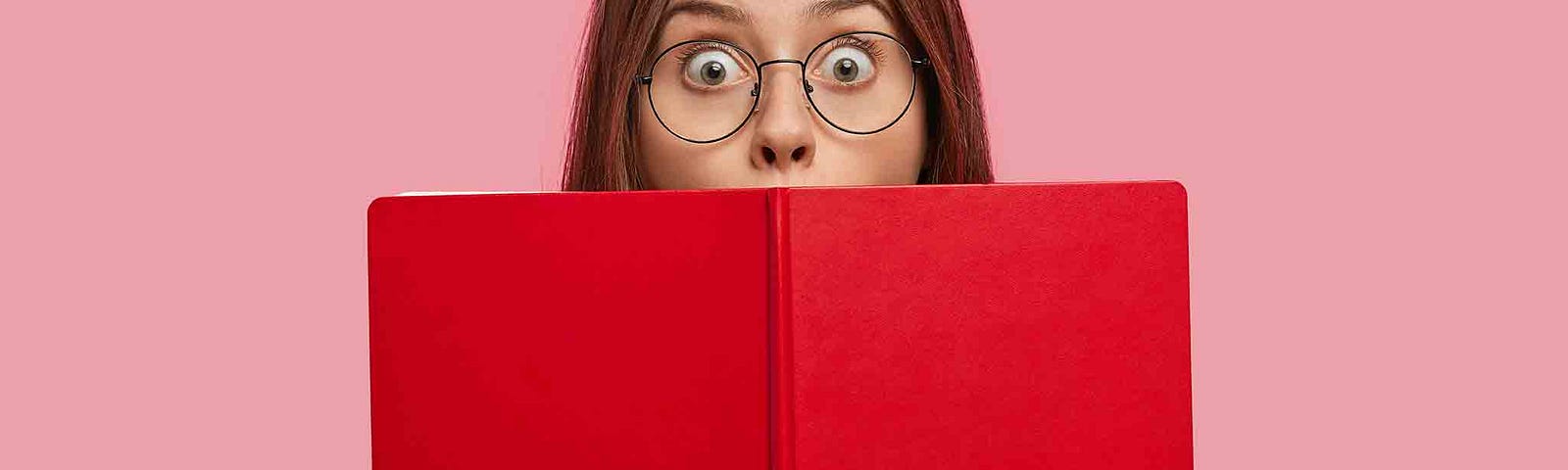 Young woman is holding a red book up to her face. It covers her mouth. Her eyes are wide with anticipation.