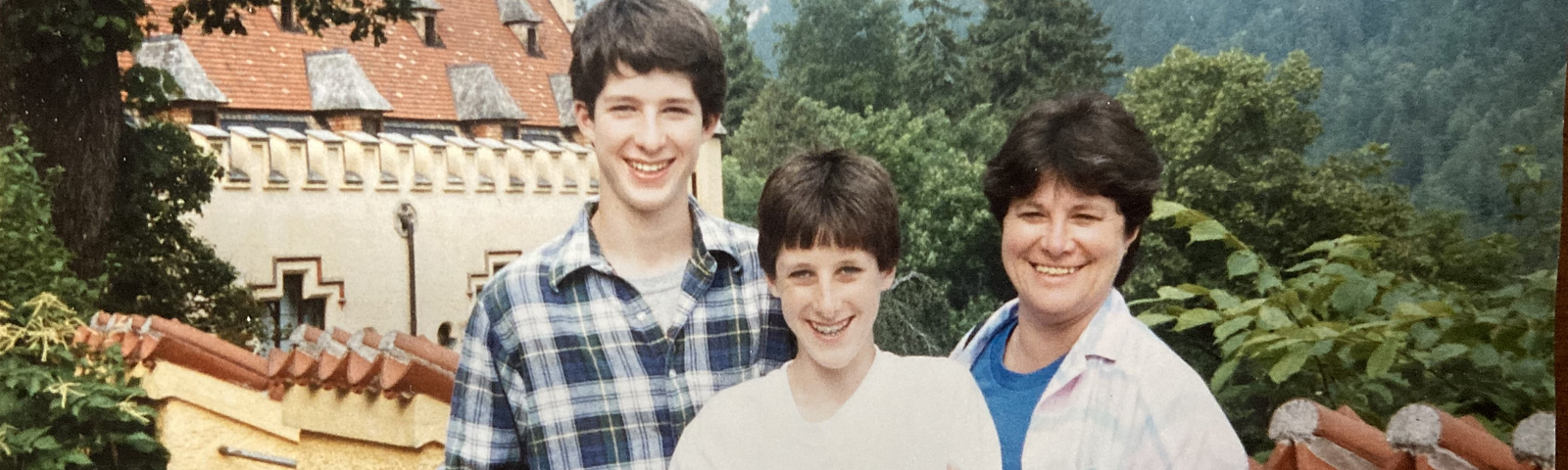 A woman and two teenage boys stand together posing for the camera. Behind them is a stone wall and behind them is part of a stone building and mountains and trees in the background.