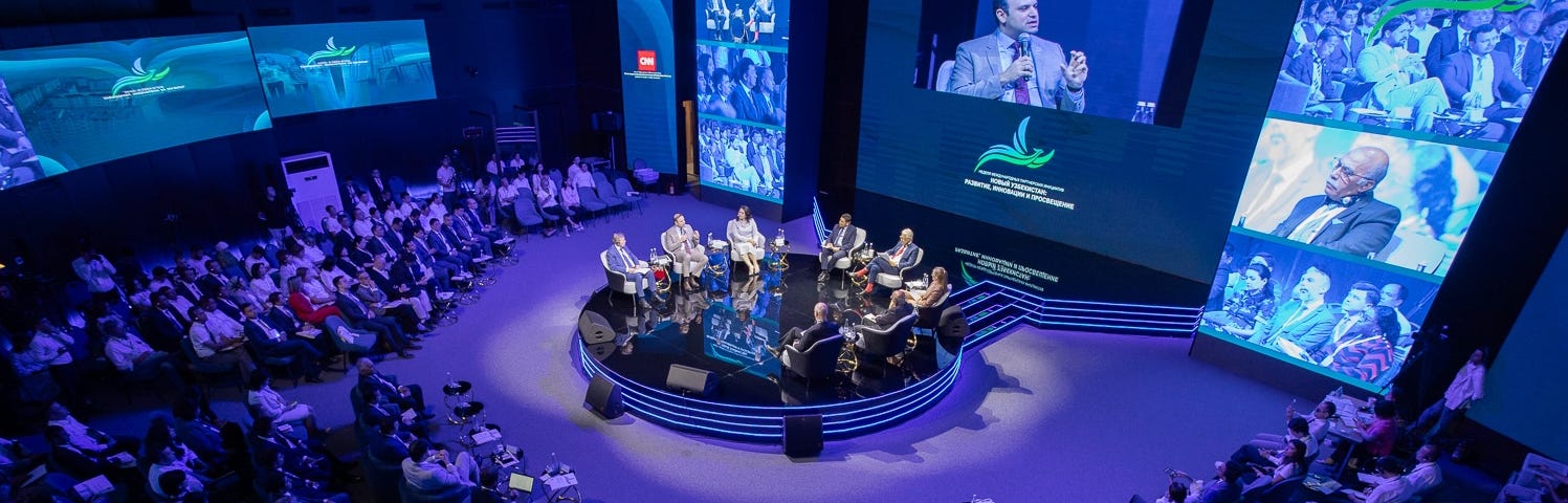 Panelists sit in the middle of a crowded room with digital screens in the background.