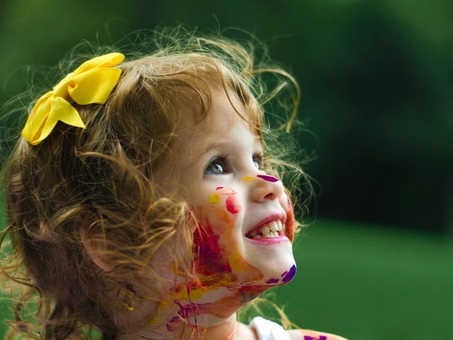 A two or three year old girl smiling at her parent or caregiver with paints on her face and dress.