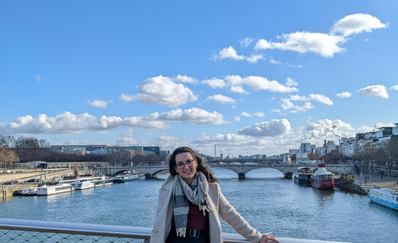 Woman wearing long coat and scarf leans against a bridge railing in front of a large river. Another bridge is in the background.