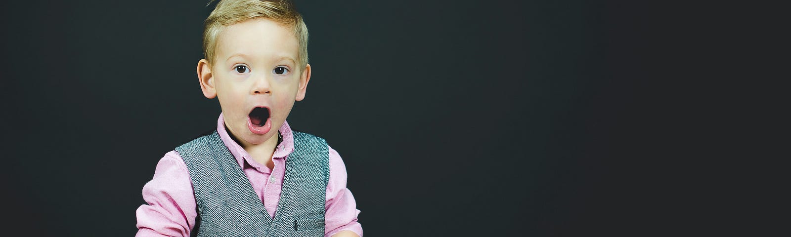Photo of a white child in jeans, pink shirt and grey waistcoat holding an open book and making a comedy surprised face