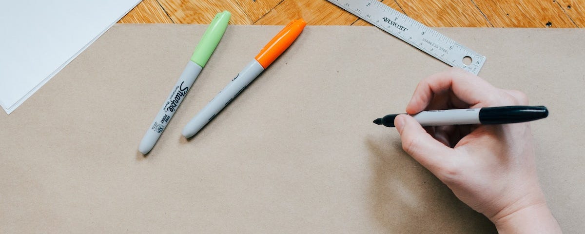 hand holding black sharpie pen on brown paper with an orange sharpie pen and a green one in upper left corner beside metal ruler.