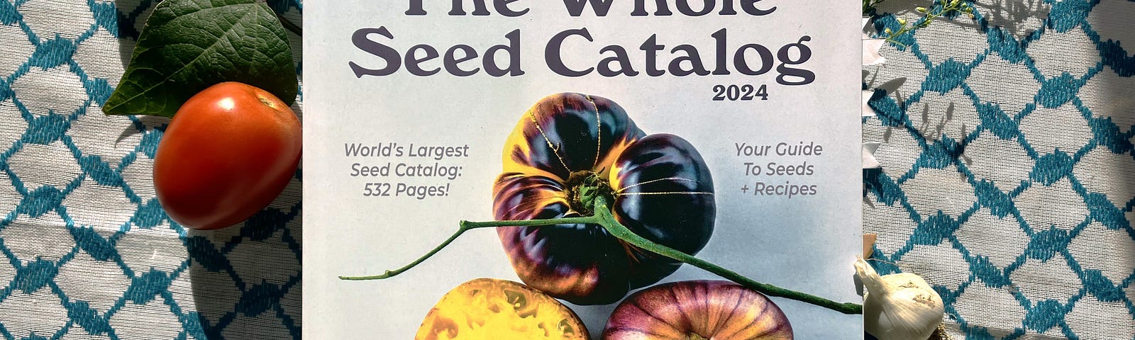 Photo of a large seed catalog titled “The Whole Seed Catalog”. The cover of the catalog boasts richly colored purple and yellow tomatoes. Leaves, garlic, and tomatoes surround the book in an flat-lay style.