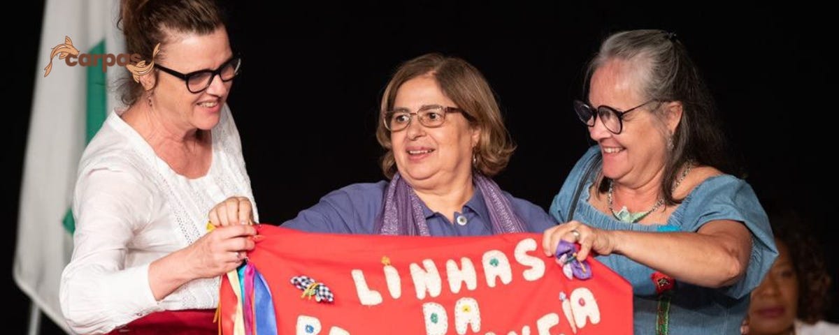 Na foto, a ministra das Mulheres Cida Gonçalves aparece ao centro segurando um tecido vermelho com os dizeres “LINHAS DA RESISTÊNCIA” em letras brancas e seis estrelas amarelas embaixo. Ao seu lado estão duas mulheres, companheiras de luta da ministra.